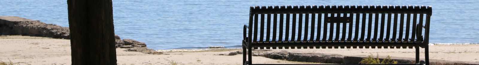 A metal bench in front of a sand beach and lake