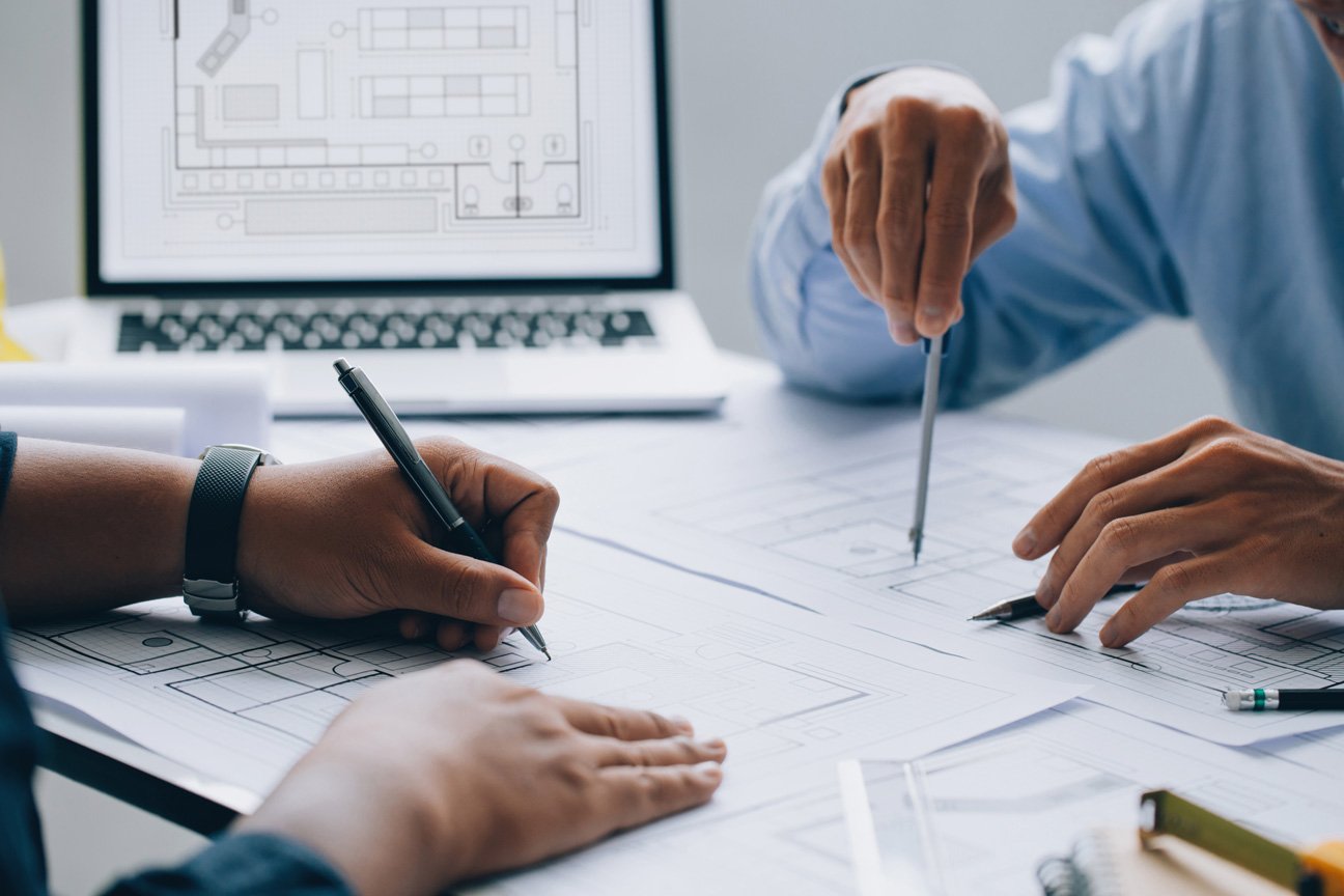 two hands working on a blueprint with a computer in the background